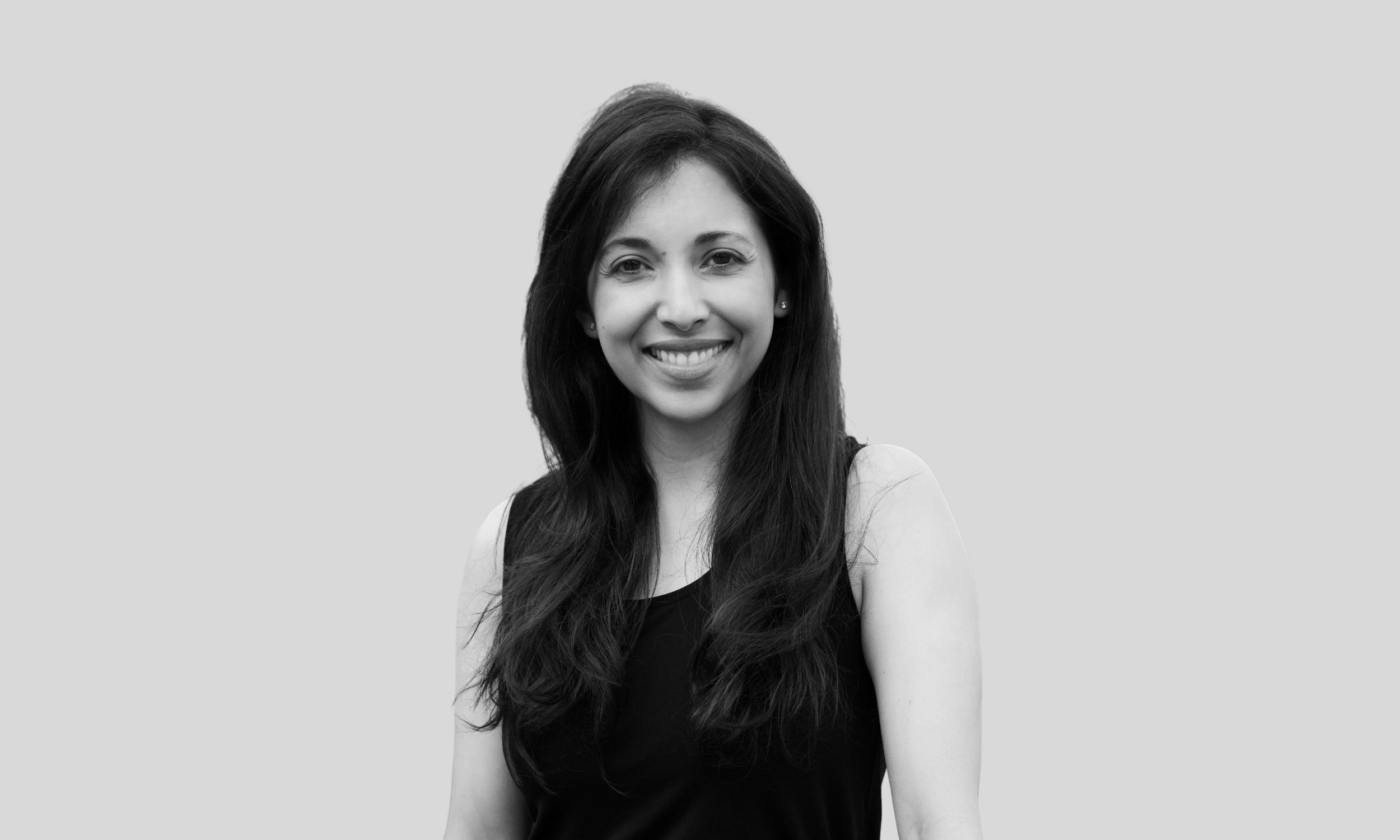 A woman faces the camera and smiles. She has long dark hair and is wearing a black sleeveless blouse.