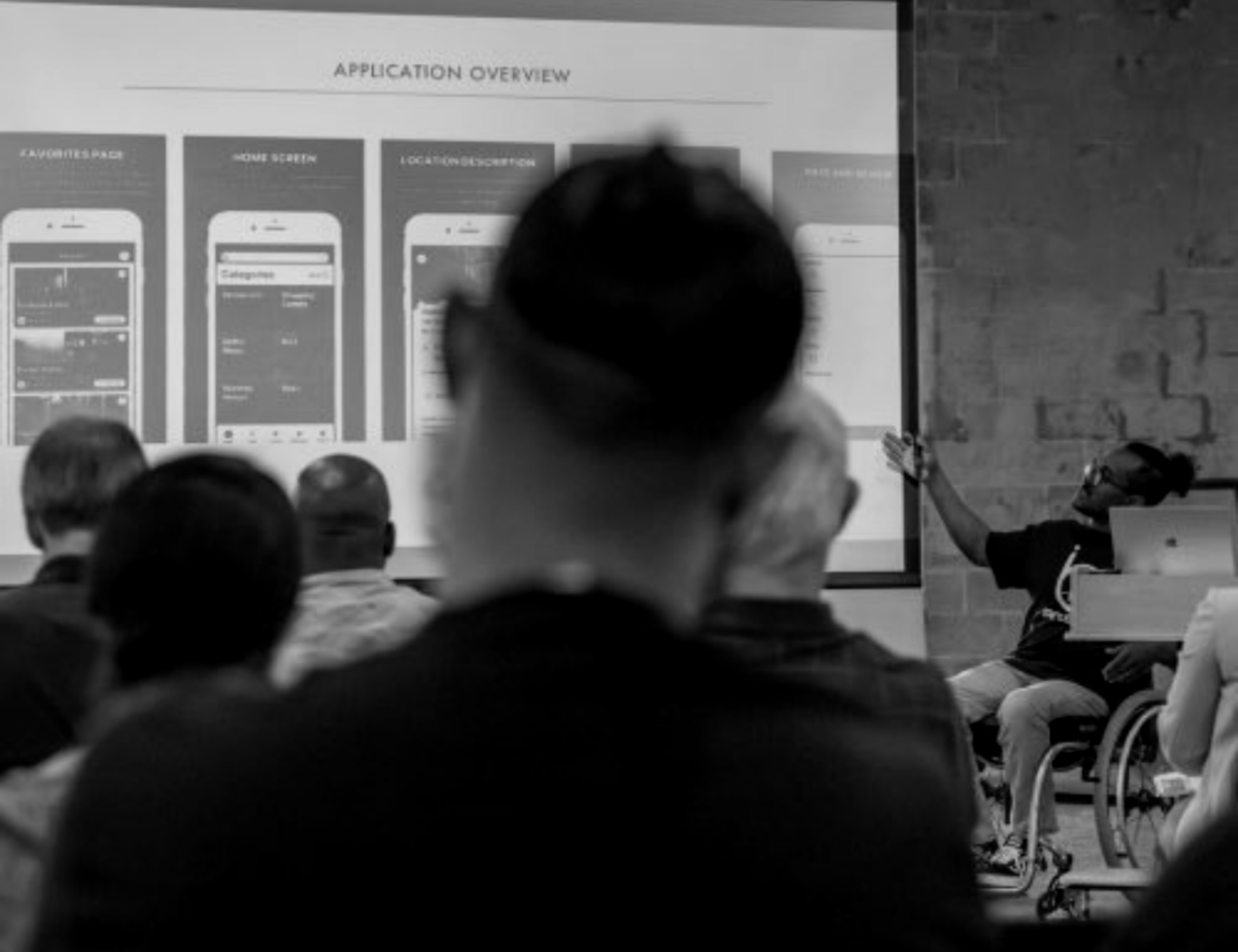 In focus is an African-American man pointing to a projector with screenshots of a mobile app on the screen. His wheelchair is partially visible. The audience is blurred in the foreground.