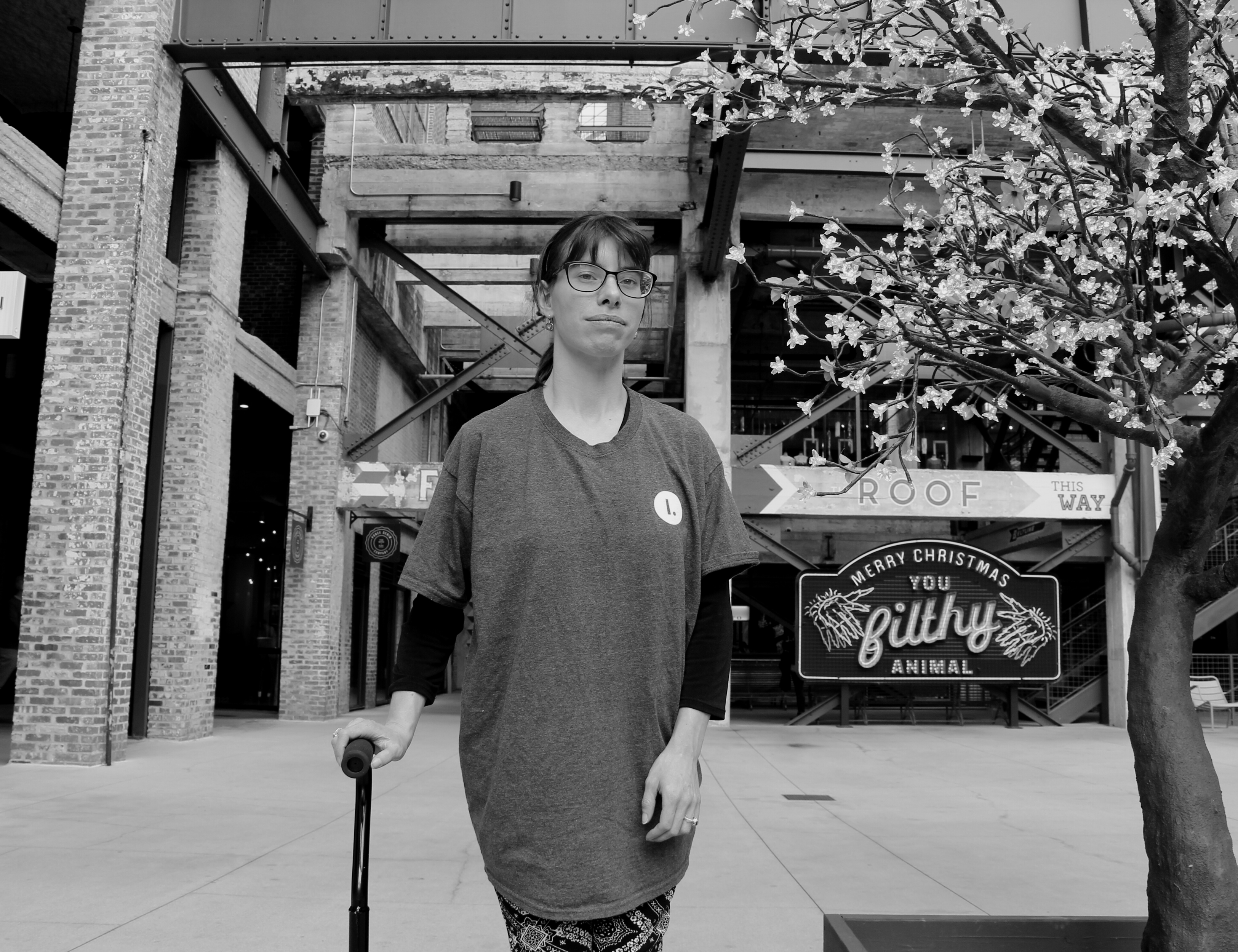 A young white woman stands facing the camera wearing an Illimitable tee shirt. Her cane is partially visible and a large brick building is visible in the background.