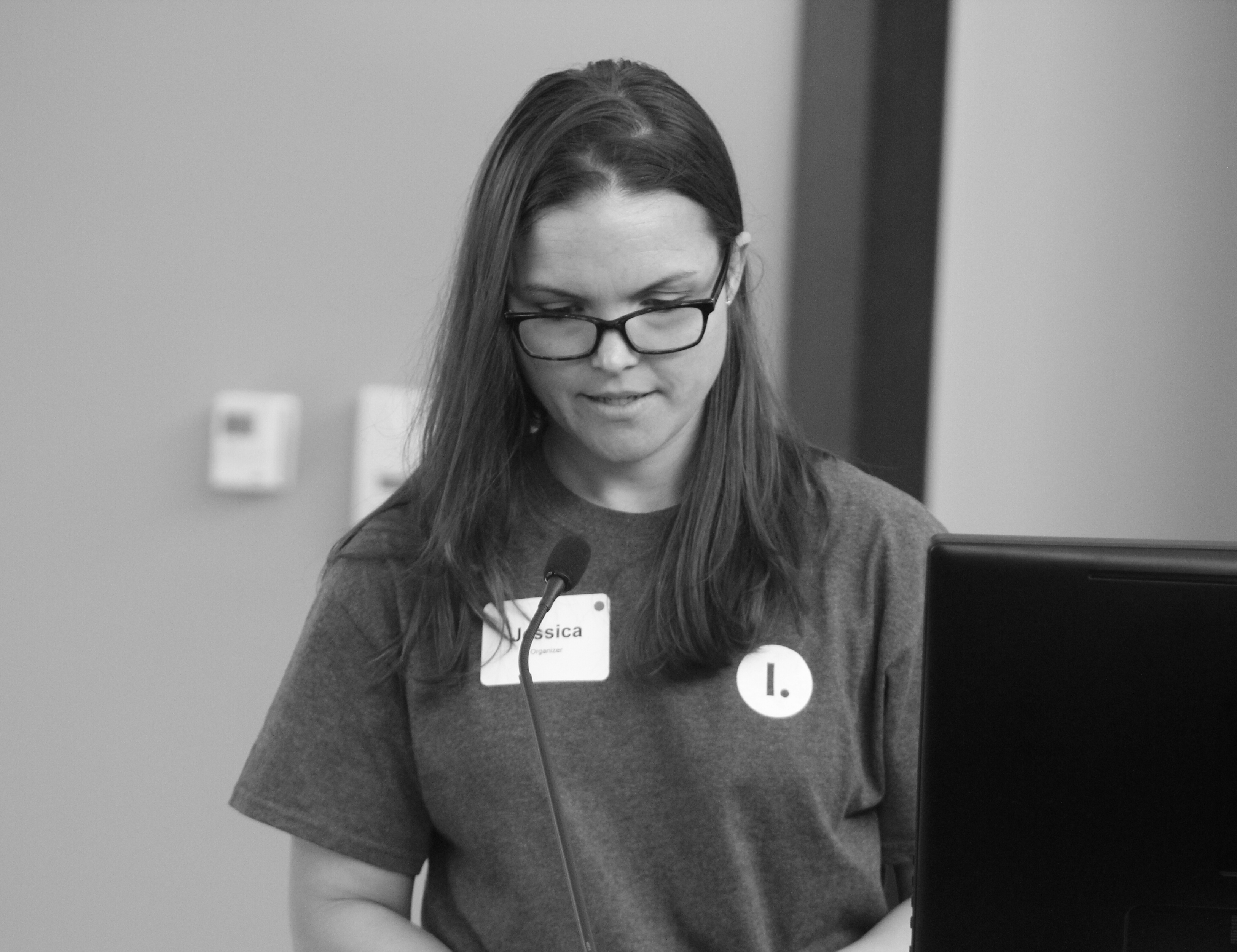 A young white woman with shoulder-length brown hair faces the camera while speaking to a crowd. She is wearing an Illimitable tee shirt and black glasses.