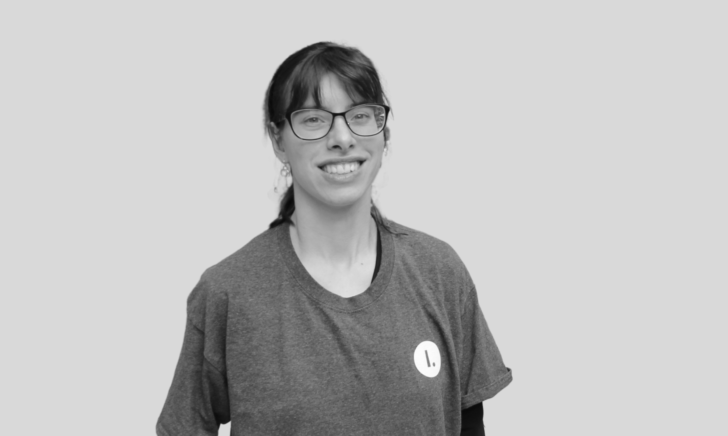 A white woman looks at the camera and smiles widely. She is wearing an Illimitable tee shirt with the logo visible, black glasses and her brown hair in a ponytail.