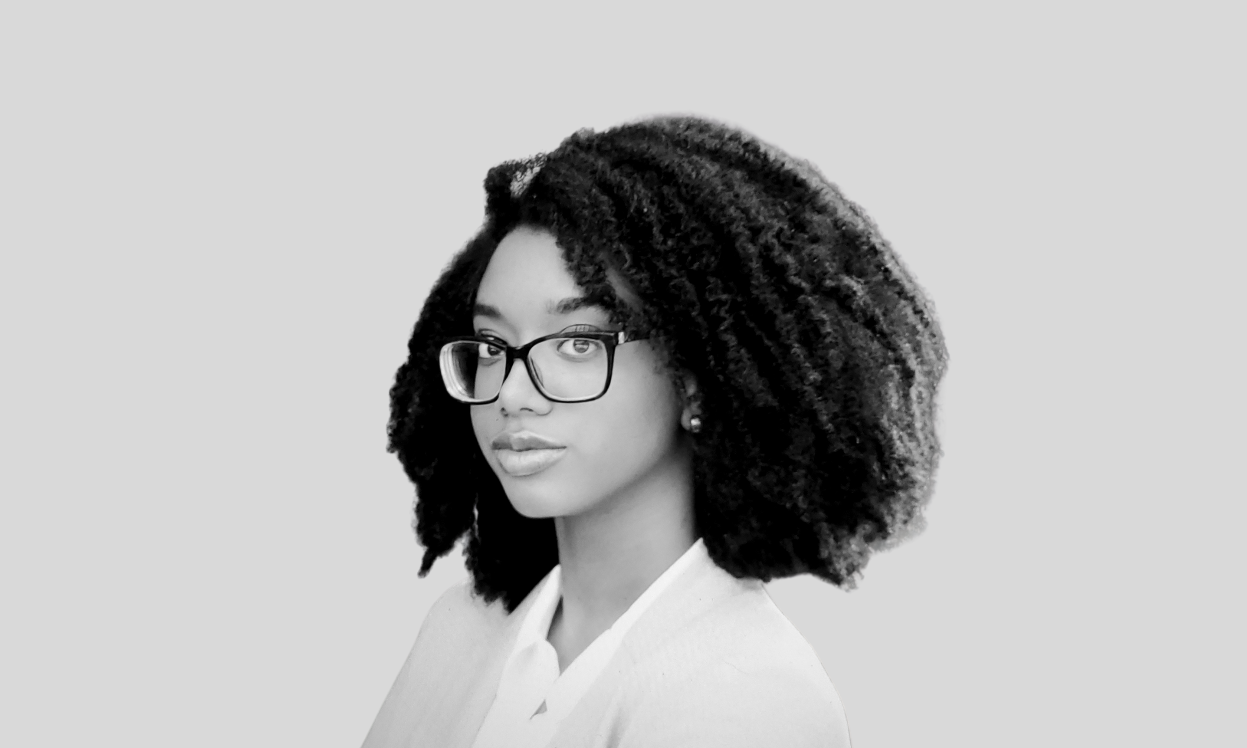 A black woman faces the camera, her brows slightly furrowed. She has course, curly black hair styled in an afro, and is wearing a long-sleeved sweater and thick black glasses.